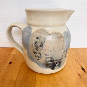 May include: A ceramic pitcher with a cream-colored body and a curved handle. The pitcher features abstract blue and gray painted designs. The spout is angled for easy pouring. The pitcher is sitting on a wooden surface.