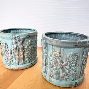 May include: Two cylindrical planters featuring a weathered teal and copper patina finish. The exterior surfaces are decorated with raised relief patterns of flowers and vertical ribbon-like columns, creating a textured appearance on both vessels.