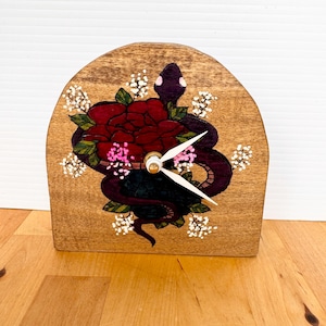 May include: An arched wooden clock featuring a hand-painted design of a dark purple snake coiled around a large red rose. The artwork includes green leaves and small white and pink floral accents. The clock has two white hands at the center.