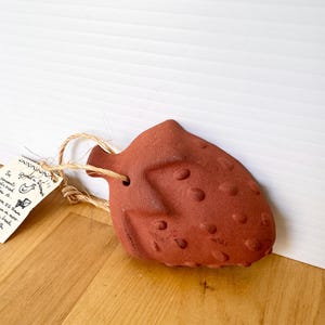 May include: A terracotta-colored, strawberry-shaped ceramic bread warmer with a textured surface featuring raised dots. A natural jute twine loop is threaded through a hole at the top. A small, rectangular paper tag with handwritten text is attached.
