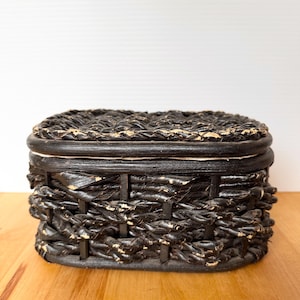 May include: A black woven basket with a lid. The basket is rectangular with a rounded top and bottom. The weave is thick and textured, with some areas showing a lighter, natural color. The basket sits on a wooden surface.