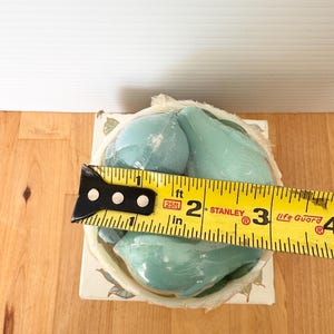 Puede incluir: Un juego de jabones en forma de pera de color azul claro, envuelto en pl&aacute;stico transparente, se encuentra en una caja decorativa. La caja est&aacute; decorada con motivos florales. Una cinta m&eacute;trica amarilla muestra que el juego de jabones mide aproximadamente 7,6 cm de ancho.