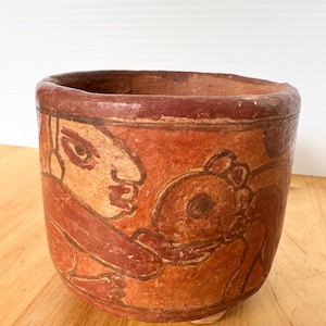 May include: A terracotta-colored ceramic pot with a rounded base and a wide rim. The pot features a reddish-brown painted design of figures. The figures are depicted in a stylized manner, with bold lines and simple shapes.