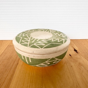 May include: A round ceramic lidded bowl featuring a matte sage green finish with etched white geometric patterns. The design consists of angular lines and triangles covering both the lid and the base, creating a cohesive visual motif across the piece.