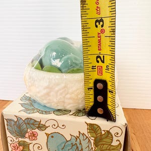 Puede incluir: Figura de p&aacute;jaro de cer&aacute;mica en un nido, empaquetada en una caja decorativa. El nido es blanco con una superficie texturizada, y el p&aacute;jaro est&aacute; envuelto en pl&aacute;stico. La caja presenta una ilustraci&oacute;n de p&aacute;jaro azul con dise&ntilde;os florales y de hojas. La altura es de aproximadamente 7,6 cm.