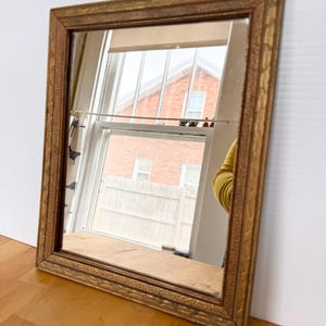 May include: A rectangular mirror with a decorative gold-colored frame. The mirror reflects a window with a view of a brick building. The frame has an ornate, textured design. The mirror is leaning against a white surface on a wooden floor.