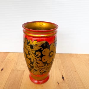 May include: A cylindrical wooden vase featuring a vibrant red base color with intricate black and gold floral patterns painted on the exterior. The interior rim and the base are finished in a solid gold tone, creating a contrast with the red body.