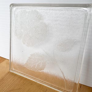 May include: A clear glass block with a floral design etched into the surface. The design features a large flower and leaves. The block is square and has a slightly frosted appearance.