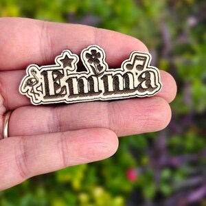 May include: Wooden name tag with the name "Emma" in a decorative font. The tag is adorned with a flamingo, star, flower, and musical note. The tag is held in a hand against a blurred green background.