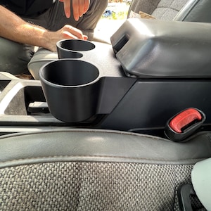 Puede incluir: Portavasos de plástico negro con dos ranuras para vasos, instalado en la consola central de un coche.