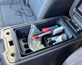 May include: A black car center console with a red button labeled "PRESS" and an open compartment containing a variety of items, including a clear plastic bag with seeds, a red and black pen, a pack of colorful markers, a pack of cigarettes, and a small white container.