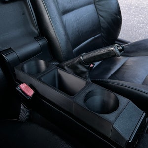 May include: Black car console cup holder with two cup holders and a storage compartment. The cup holder is installed in a black leather car interior. The car's emergency brake is visible.