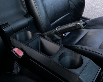 May include: Black car console cup holder with two cup holders and a storage compartment. The cup holder is installed in a black leather car interior. The car's emergency brake is visible.