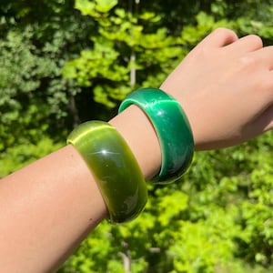 Pode incluir: Duas pulseiras verdes são usadas em um braço. As pulseiras são de cor verde lisa e brilhante. O fundo é uma vista desfocada da folhagem verde.