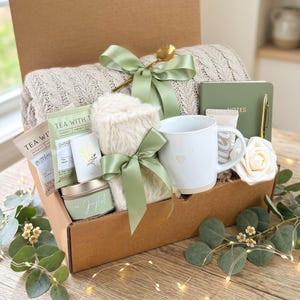 May include: A gift box filled with a beige knit blanket, a white mug with a heart, tea packets, a candle, a notebook, and a faux fur item, all tied with sage green ribbons. A gold spoon and a white rose are also included.