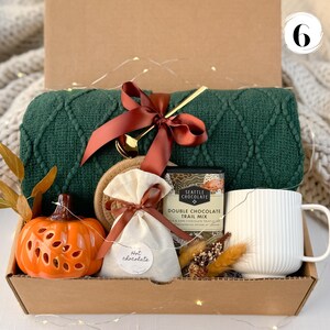 May include: A gift box filled with a green knit blanket, a ceramic pumpkin, a white mug, a bag of hot chocolate, and a chocolate bar. The box is tied with a brown ribbon and includes a gold spoon and a coaster. The overall theme is cozy and autumnal.