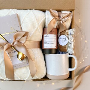 May include: A gift box filled with a white blanket, a mug, chocolate, tea, and sea salt caramels. The items are tied with gold satin ribbons. The mug is white with a tan base and speckled design. The chocolate bar is labeled "moonstruck".