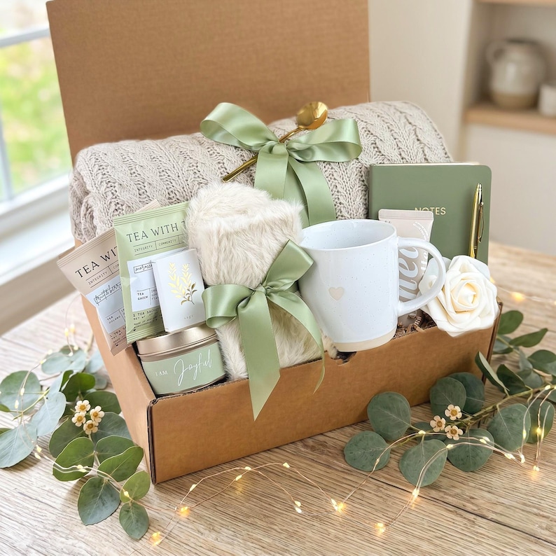May include: A gift box filled with a beige knit blanket, a white mug with a heart, tea packets, a candle, a notebook, and a faux fur item, all tied with sage green ribbons. A gold spoon and a white rose are also included.