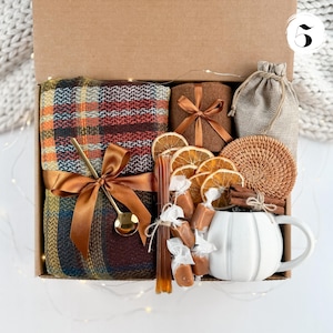May include: A gift box filled with cozy items. It includes a plaid blanket, a brown candle, a small burlap bag, a woven coaster, dried orange slices, wrapped candies, a honey stick, a gold spoon, a cinnamon stick, and a white mug.