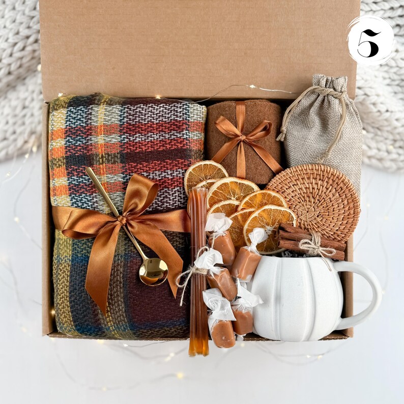 May include: A gift box filled with cozy items. It includes a plaid blanket, a brown candle, a small burlap bag, a woven coaster, dried orange slices, wrapped candies, a honey stick, a gold spoon, a cinnamon stick, and a white mug.