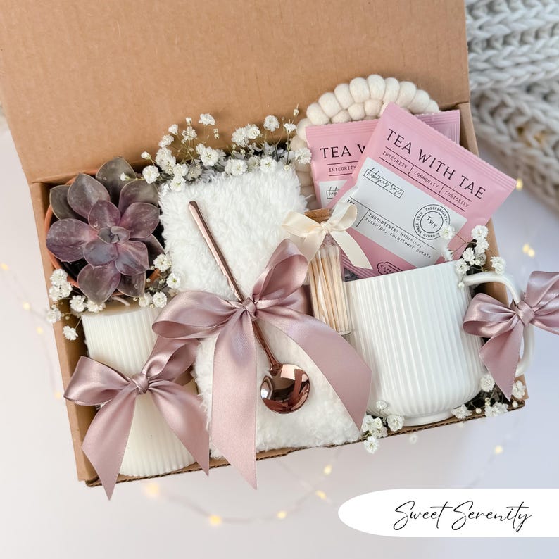 May include: A gift box filled with a succulent, tea packets, a white mug, a fluffy white cloth, a candle, and a spoon. The items are tied with pink satin bows and surrounded by baby's breath. The box is labeled "Sweet Serenity."