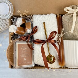 May include: A gift box containing a selection of items. It features a glass jar with wrapped caramels, a white fluffy towel tied with a brown ribbon, a gold spoon, a candle, a burlap bag, and a white mug. The candle reads "Pumpkin Clove".