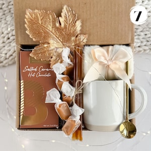 May include: A gift box containing a white mug, gold spoon, salted caramel hot chocolate, wrapped caramels, and a decorative gold leaf. The box also includes a small, fluffy blanket tied with a bow.