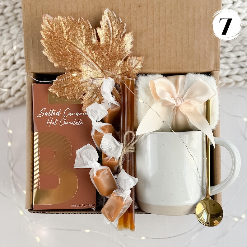May include: A gift box containing a white mug, gold spoon, salted caramel hot chocolate, wrapped caramels, and a decorative gold leaf. The box also includes a small, fluffy blanket tied with a bow.