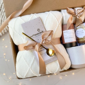 May include: A gift box filled with a cream-colored blanket, a bar of Moonstruck milk chocolate, a gold spoon, tea, cookies, and a white mug. The items are tied with a light brown ribbon.
