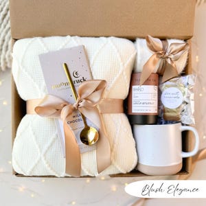 May include: A gift box with a cream-colored waffle-knit blanket, a chocolate bar with a gold spoon, a mug, tea, and sea salt caramels. The items are tied with a blush-colored ribbon. The text "Blush Elegance" is at the bottom.