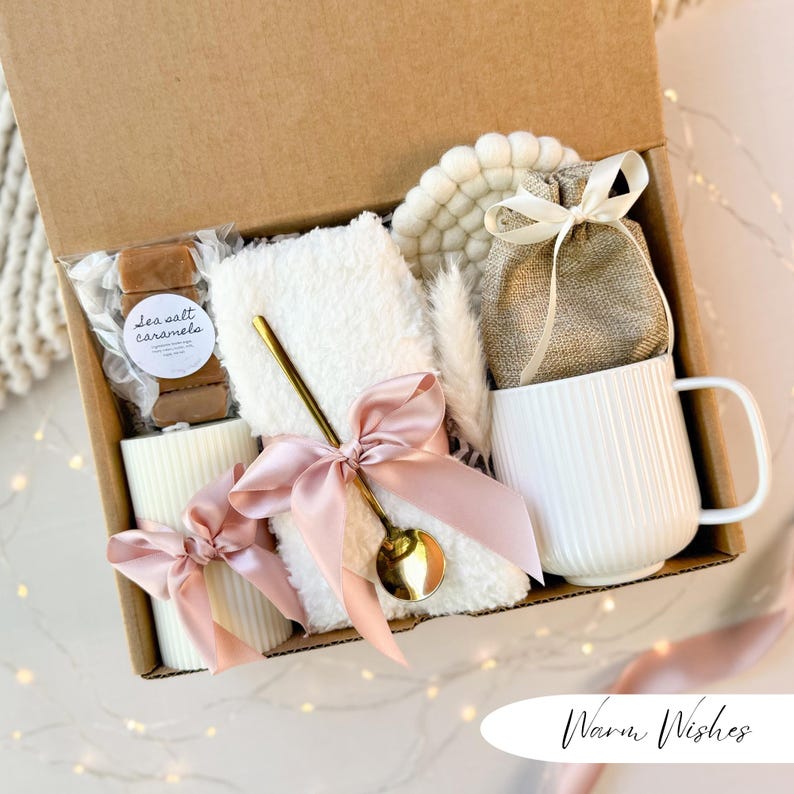 May include: A gift box filled with a white mug, a small white towel, a gold spoon, sea salt caramels, a candle, and a coaster. The items are tied with pink ribbons. The box is brown and has the words "Warm Wishes" on the bottom right.