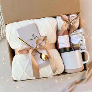 May include: A gift box filled with a cream-colored waffle-knit blanket, a bar of Moonstruck milk chocolate, a gold spoon, a mug, tea, and cookies. The items are tied with a satin ribbon. The box is brown.