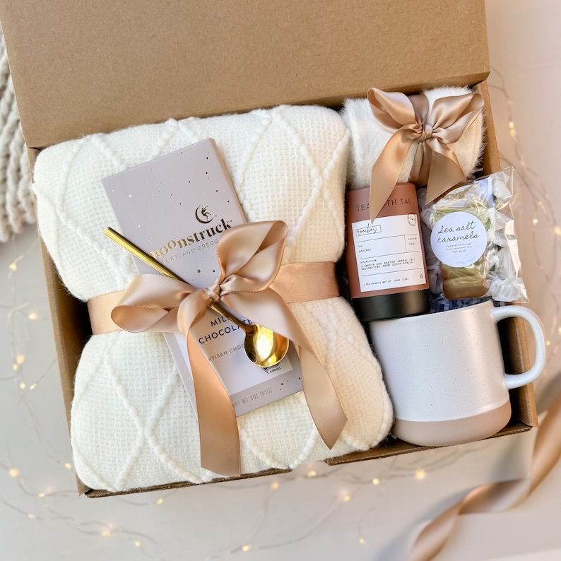 May include: A gift box filled with a cream-colored waffle-knit blanket, a bar of Moonstruck milk chocolate, a gold spoon, a mug, tea, and cookies. The items are tied with a satin ribbon. The box is brown.