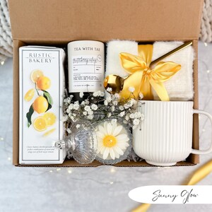 May include: A gift box filled with a white mug, a candle, a box of shortbread cookies, a small white blanket tied with a yellow ribbon, and a gold spoon. The box is decorated with white flowers and a daisy-shaped candle.