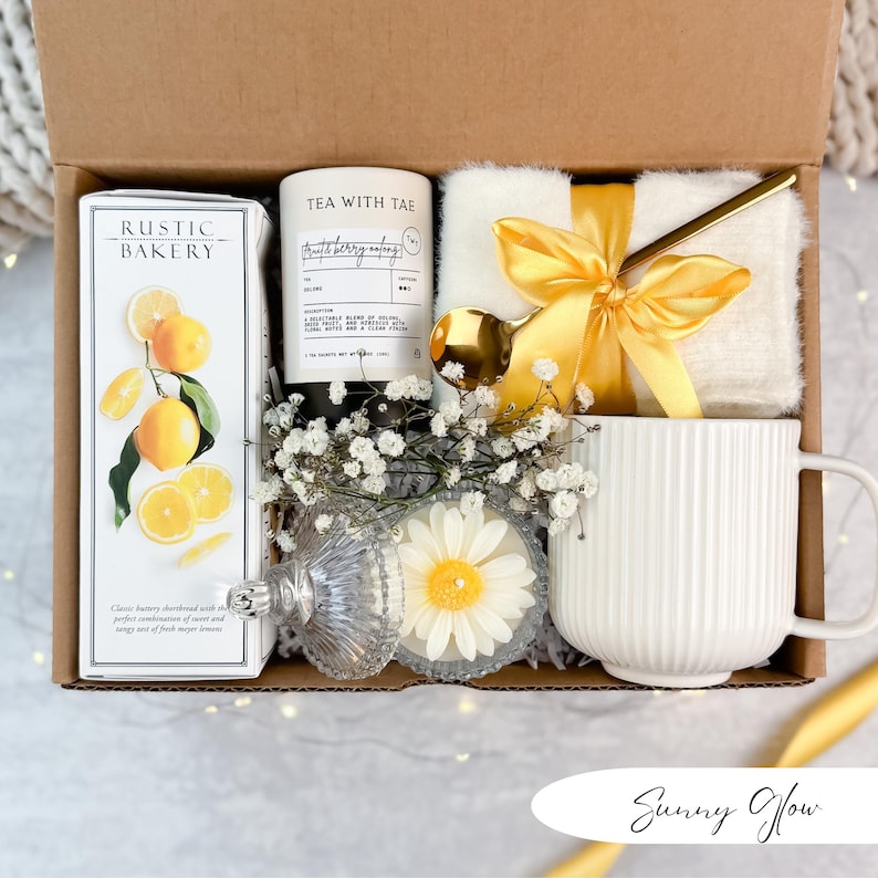 May include: A gift box filled with a white mug, a candle, a box of shortbread cookies, a small white blanket tied with a yellow ribbon, and a gold spoon. The box is decorated with white flowers and a daisy-shaped candle.