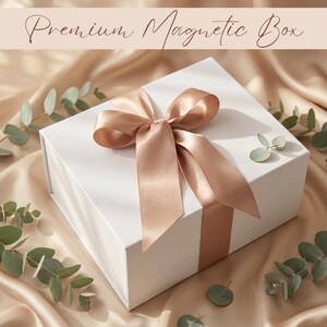 May include: A white magnetic gift box with a satin ribbon tied in a bow. The ribbon and bow are a light peach color. The box is surrounded by eucalyptus leaves and sits on a satin fabric. The text "Premium Magnetic Box" is at the top.