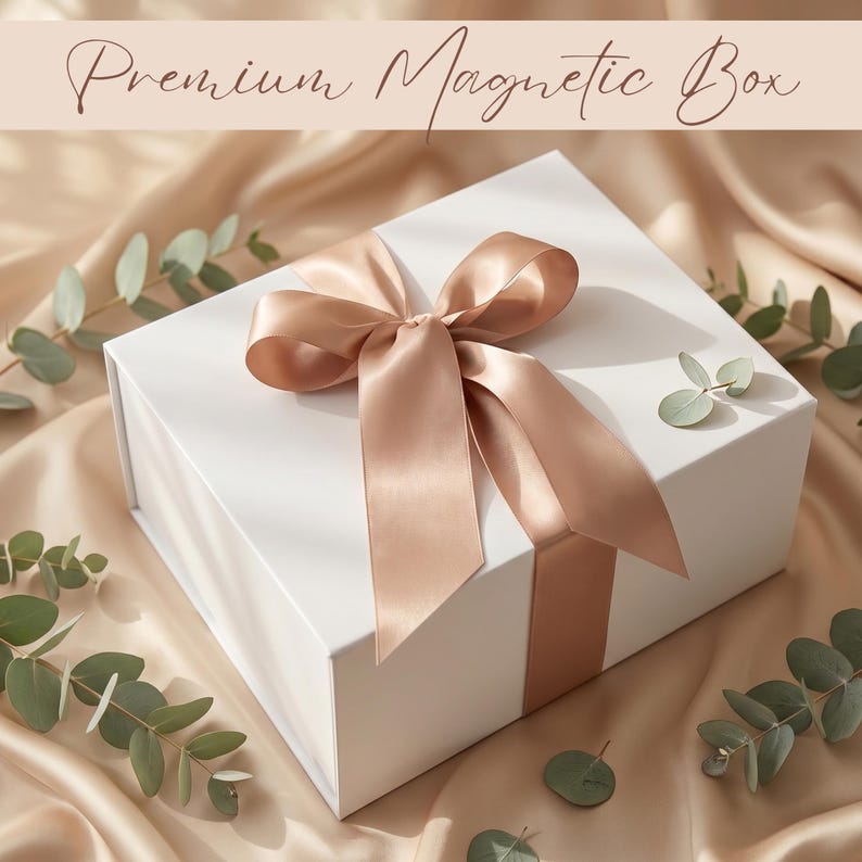 May include: A white magnetic gift box with a satin ribbon tied in a bow. The ribbon and bow are a light peach color. The box is surrounded by eucalyptus leaves and sits on a satin fabric. The text "Premium Magnetic Box" is at the top.