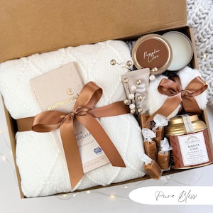 May include: A gift box filled with a white blanket, chocolate bar, candle, tea, and other treats. The box is tied with a brown ribbon. The box includes a candle labeled "Pumpkin Clove". The text "Pure Bliss" is at the bottom.