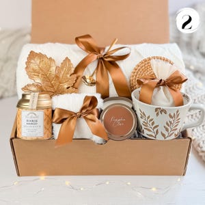 May include: A gift box filled with a white blanket, a mug with a leaf design, a candle, tea, and a decorative leaf. The items are tied with brown satin ribbons. A small gold spoon rests on the blanket.