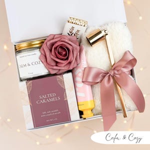 May include: A white gift box filled with self-care items. Includes a candle, matches, a rose, hand cream, a box of salted caramels, and a gold candle snuffer with a pink ribbon. The candle reads "CALM & COZY".