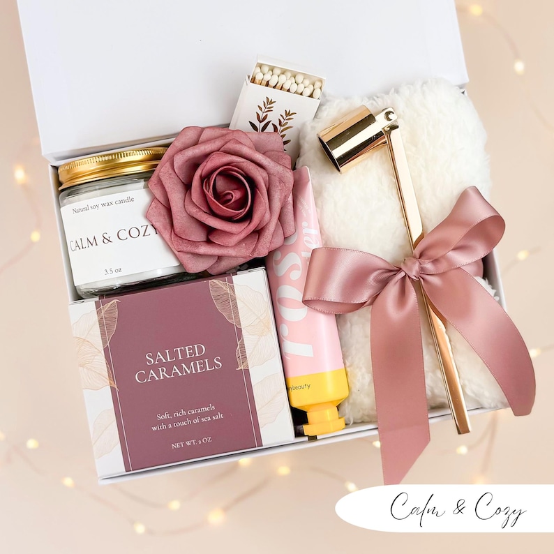 May include: A white gift box filled with self-care items. Includes a candle, matches, a rose, hand cream, a box of salted caramels, and a gold candle snuffer with a pink ribbon. The candle reads "CALM & COZY".