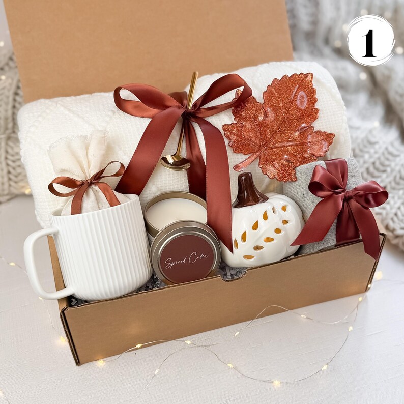 May include: A gift box filled with fall-themed items. Includes a white ribbed mug, a candle, a decorative pumpkin, a blanket, and a decorative leaf. The items are tied with brown ribbons. The box is brown.