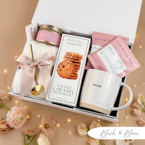 May include: A gift box filled with a white mug with "mom" printed on it, cookies, tea packets, a candle, a gold spoon, and a fluffy white blanket tied with a pink ribbon. The box is labeled "Blush & Bloom".