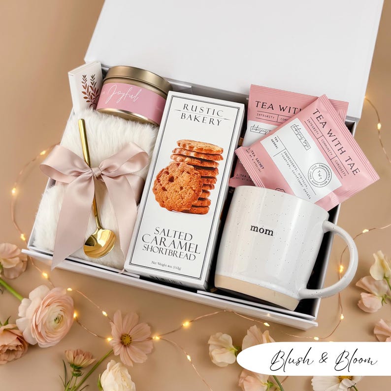 May include: A gift box filled with a white mug with "mom" printed on it, cookies, tea packets, a candle, a gold spoon, and a fluffy white blanket tied with a pink ribbon. The box is labeled "Blush & Bloom".