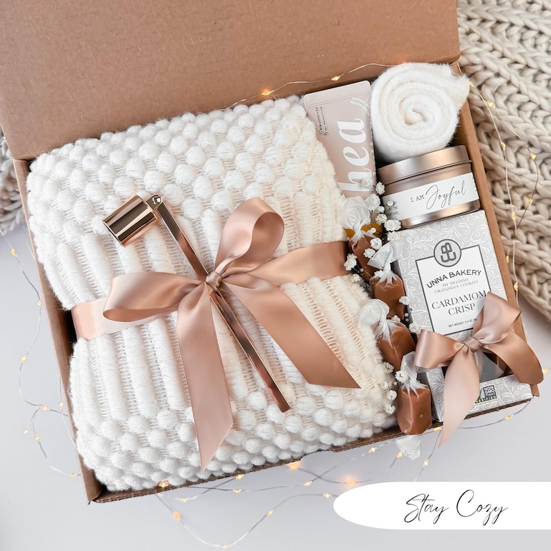 May include: A gift box filled with cozy items. It includes a white textured blanket tied with a peach satin ribbon, a candle, a tube of lotion, and a box of cardamom crisp cookies. The box is decorated with fairy lights and the words "Stay Cozy".