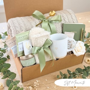 May include: A gift box filled with self-care items. Includes a beige knit blanket, a white mug with a heart, tea packets, a candle, a notebook, a pen, a gold spoon, and a faux fur item, all tied with sage green ribbons. The box is brown.