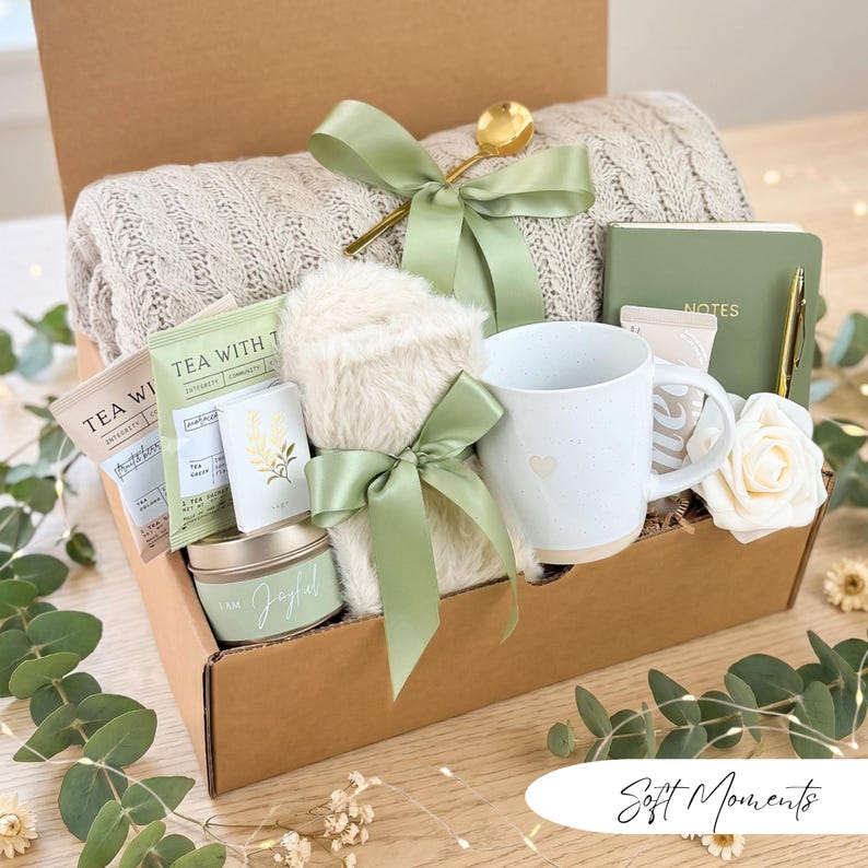 May include: A gift box filled with self-care items. Includes a beige knit blanket, a white mug with a heart, tea packets, a candle, a notebook, a pen, a gold spoon, and a faux fur item, all tied with sage green ribbons. The box is brown.