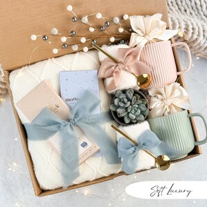May include: A gift box filled with a cream-colored cable-knit blanket, two ribbed ceramic mugs, a small succulent, and two small notebooks. The box is decorated with gold spoons, silk bows, and decorative sprigs.