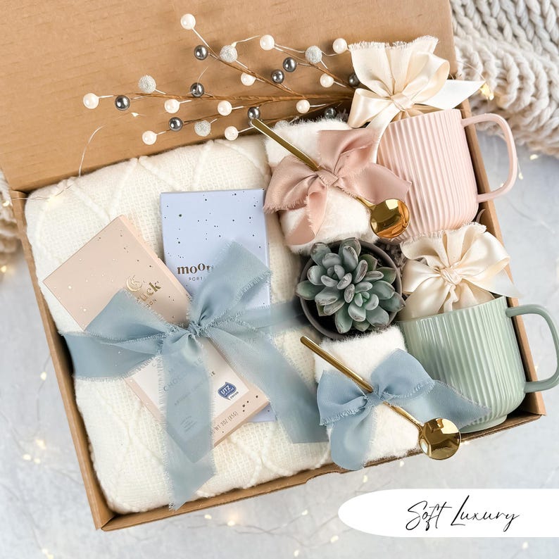 May include: A gift box filled with a cream-colored cable-knit blanket, two ribbed ceramic mugs, a small succulent, and two small notebooks. The box is decorated with gold spoons, silk bows, and decorative sprigs.