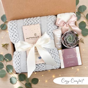 May include: A gift box filled with a gray knit blanket, chocolate bar, hand cream, a small succulent, a white washcloth with a pink bow, and a box of salted caramels. The box is tied with a white ribbon. The text "Cozy Comfort" is at the bottom.